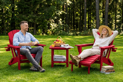 A man and a woman relax in red Adirondack chairs on a green lawn, with a small red side table between them holding a fruit basket and a rolled towel. Trees fill the background.