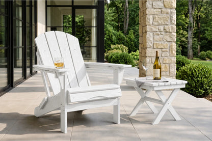 A white outdoor Adirondack chair with a matching side table on a patio, with a glass of whiskey in the chair's cup holder and a wine bottle on the table. The background shows a stone pillar, glass patio doors, and a lush green garden.
