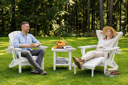 A man and a woman relax in white outdoor chairs on a grassy lawn with a small table between them, set against a backdrop of a dense green forest.