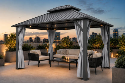 A dark gray hardtop gazebo with netting and curtains, furnished with a beige sofa, black armchairs, and a coffee table on a rooftop terrace at dusk with a city skyline in the background.