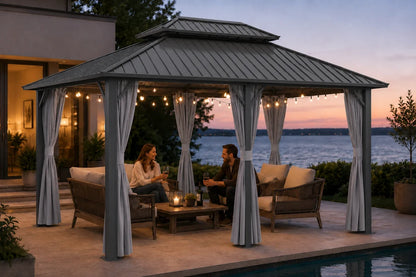 A dark gray hardtop gazebo with netting and curtains, set on a poolside patio at dusk. Two people are seated underneath on a patio sofa and coffee table, with a house, lake, and sunset sky in the background.