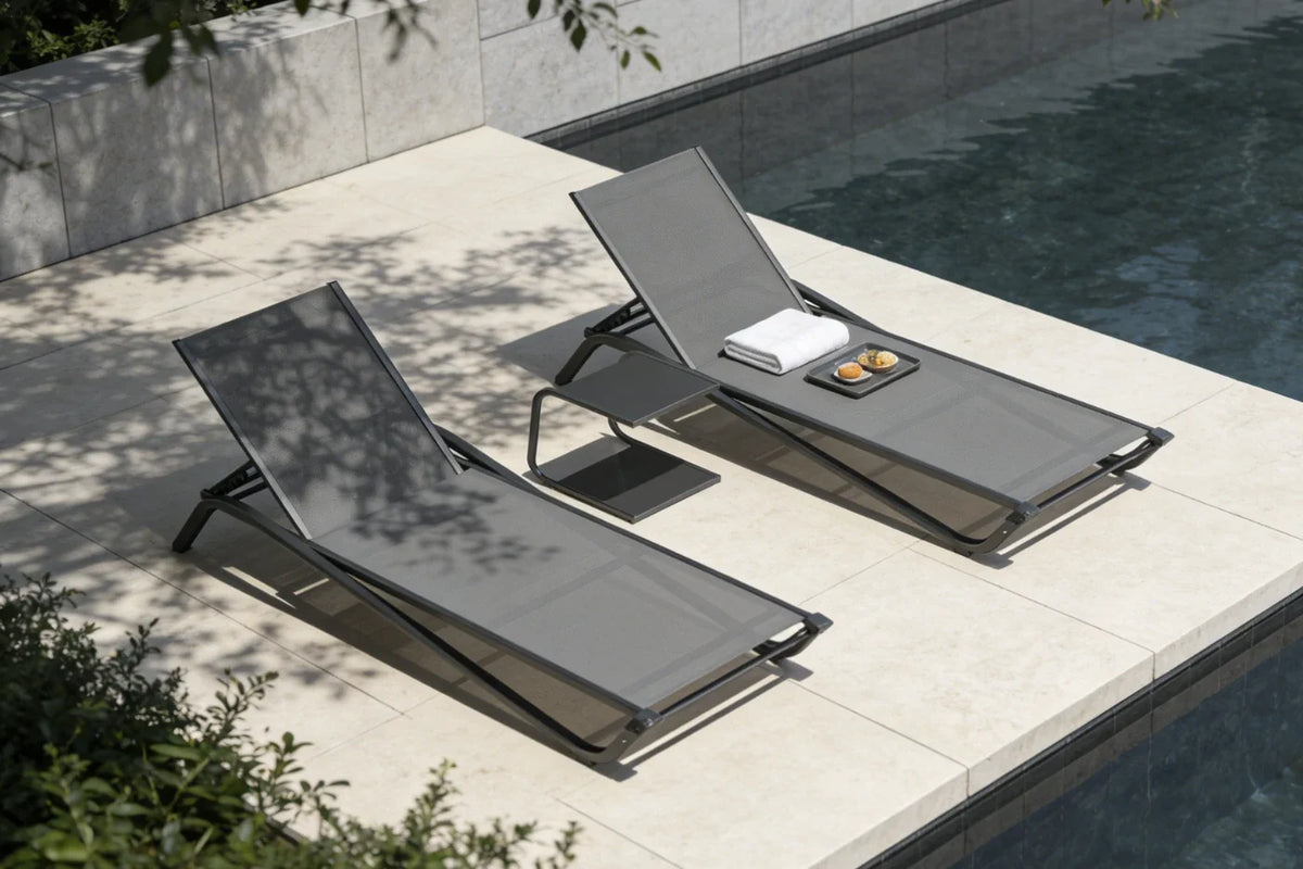 Two gray lounge chairs by a poolside with a book and a glass on a stone deck.