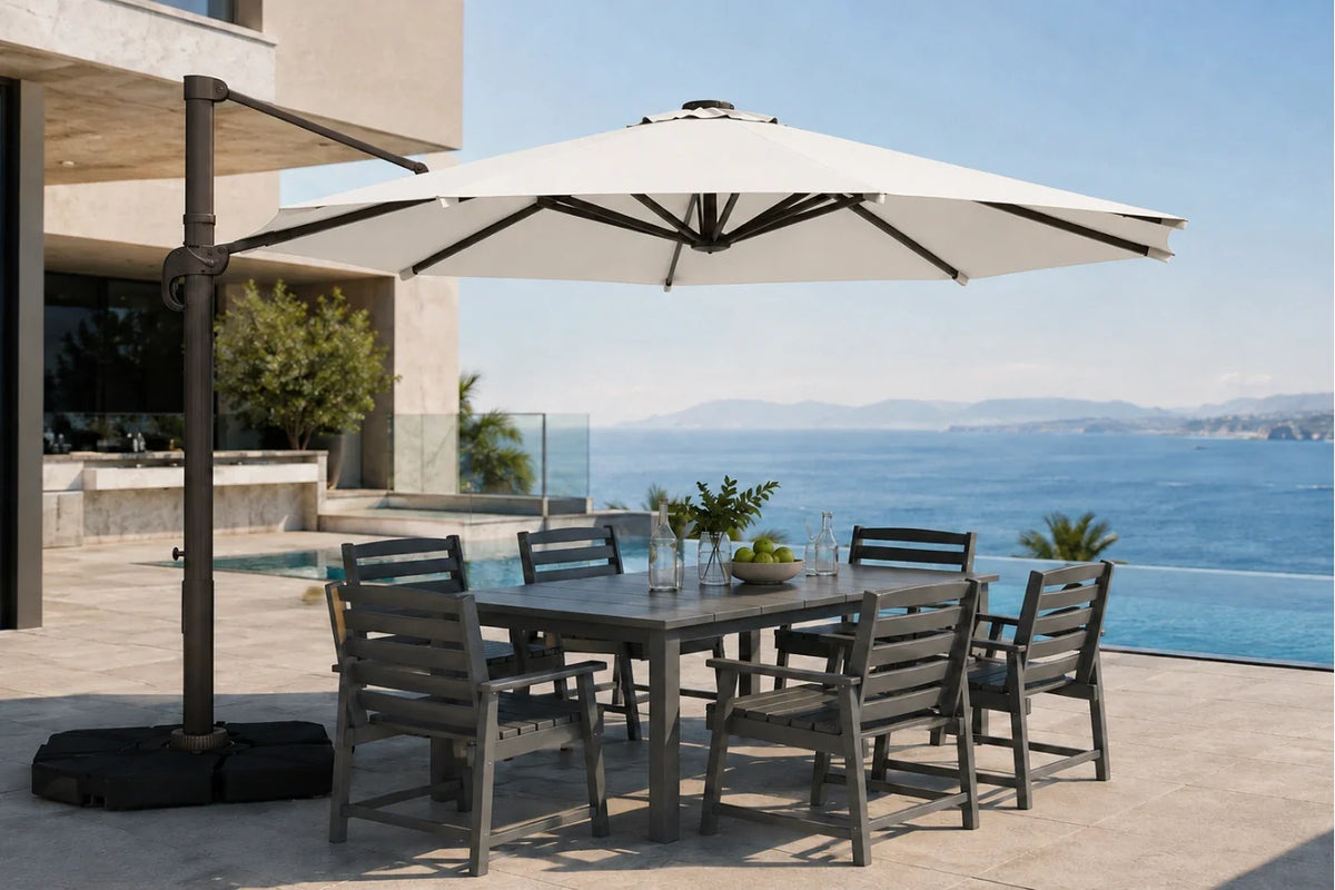 A large outdoor patio umbrella with a dark metal cantilever arm and black base, shading a rectangular gray dining table and six matching slatted chairs on a sunlit terrace overlooking a blue ocean and pool.