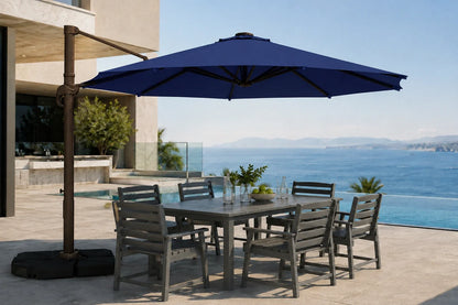 A large outdoor patio umbrella with a dark metal cantilever arm and black base, shading a rectangular gray dining table and six matching slatted chairs on a sunlit terrace overlooking a blue ocean and pool.