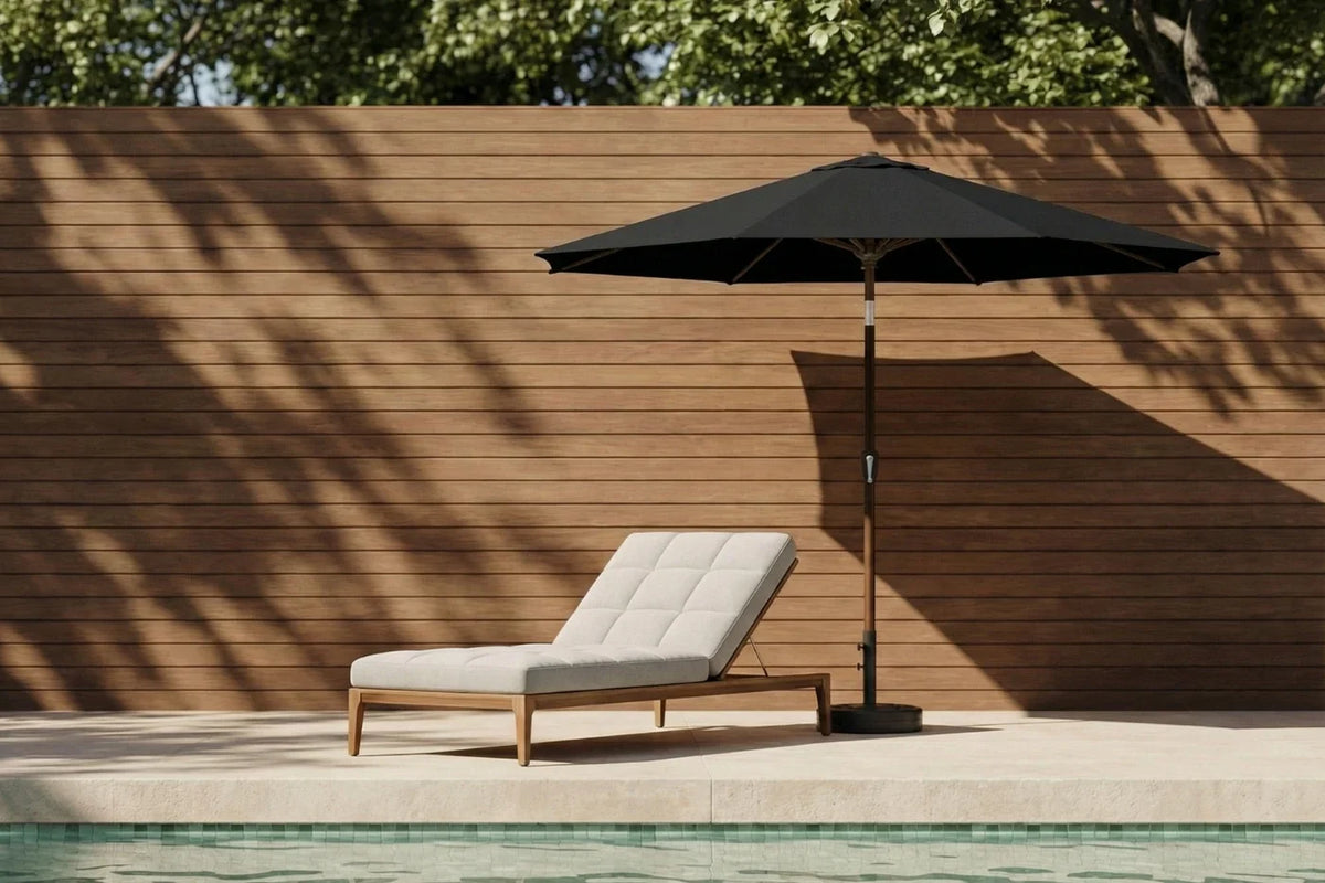 Chaise lounge with a black umbrella by a poolside against a wooden wall.