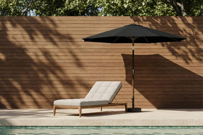 Chaise lounge with a black umbrella by a poolside against a wooden wall.