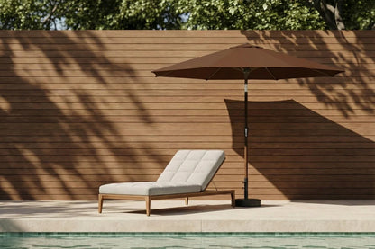 Chaise lounge with a brown umbrella by a poolside against a wooden wall.