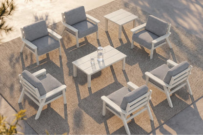 An outdoor patio conversation set with six white-framed armchairs with gray cushions arranged around one matching rectangular wooden coffee table and one matching side table on a woven rug.