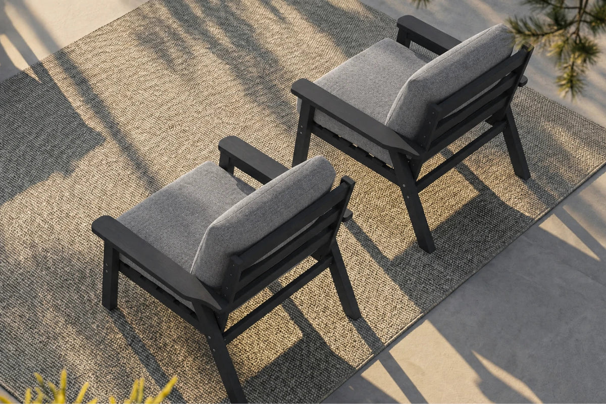 Two outdoor armchairs with black frames and gray cushions, arranged on a textured beige rug in a patio setting with natural shadows.
