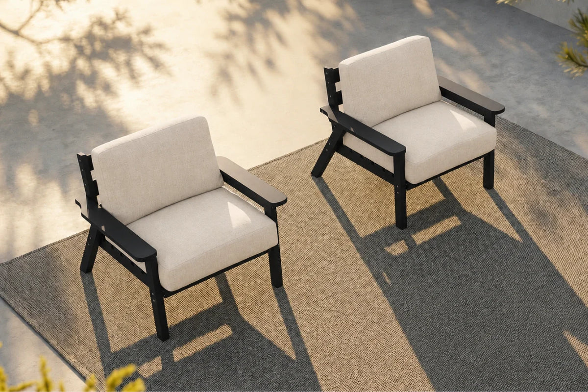 Two outdoor armchairs with black metal frames and creamy white cushions, positioned on a textured gray patio under warm sunlight.