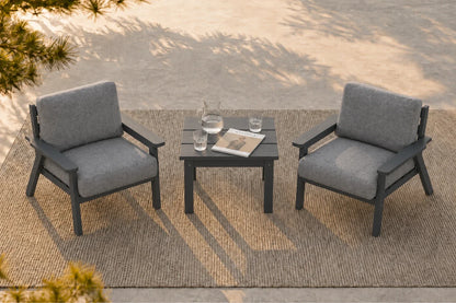 Two gray outdoor armchairs with dark gray frames arranged around a small square dark gray side table on a woven rug, photographed in a real patio setting with sunlight and tree shadows.