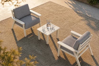Two white outdoor armchairs with gray cushions and a matching small square side table, arranged on a textured patio rug in a sunlit patio setting.