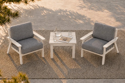 Two white outdoor armchairs with gray cushions are arranged around a small white side table on a woven rug, photographed from above in a patio setting with dappled sunlight.