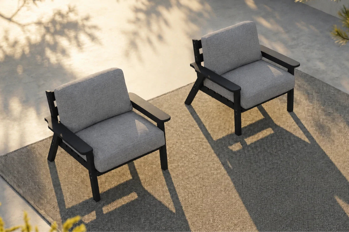Two outdoor armchairs with gray textured cushions and black frames, positioned on a textured gray patio rug with warm sunlight and leaf shadows.