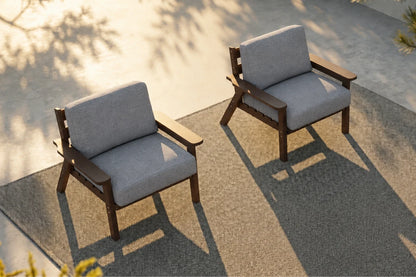 Two modern outdoor armchairs with gray textured cushions and brown wooden frames, positioned on a textured concrete patio with warm sunlight and leaf shadows.