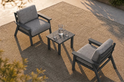 Two gray cushioned outdoor armchairs with a small square dark side table, arranged on a woven beige rug in a patio setting.