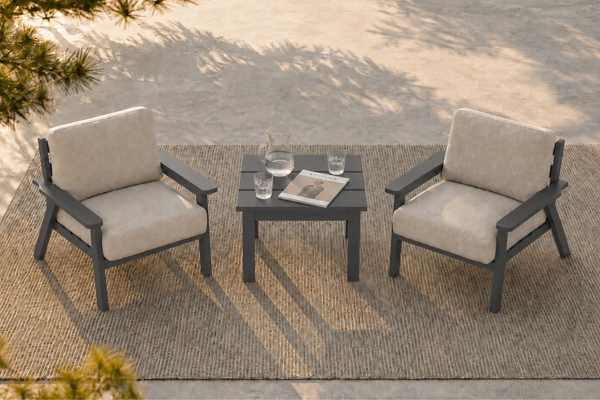 Two outdoor armchairs with creamy white cushions and dark gray frames, arranged on a woven rug around a small square dark gray side table with a glass decanter and two glasses, set on a concrete patio.