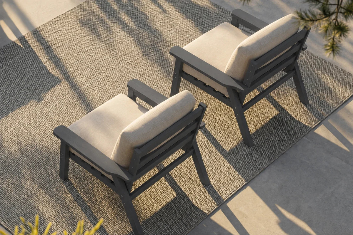 Two outdoor armchairs with dark gray frames and creamy white cushions, arranged on a textured beige rug over a concrete patio with dappled sunlight.