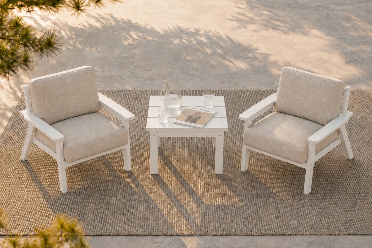 A 3-piece outdoor patio conversation set featuring two white armchairs with creamy white cushions and a matching square white side table, arranged on a woven jute rug over a concrete patio.