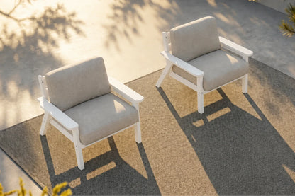 Two modern outdoor armchairs with white frames and creamy white cushions, placed on a textured patio rug in warm sunlight.